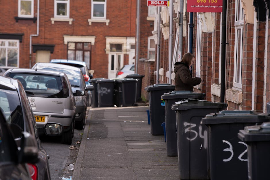 A row of large black wheelie bins with closed lids lined up along the sidewalk outside red-brick terraced houses in an urban residential area. The bins are positioned on the right side of the image, with some marked with white spray-painted numbers. A woman dressed in a dark coat is standing beside one of the bins, appearing to be in the process of accessing or closing it. The street is narrow, with parked cars on the left, including a silver hatchback in the foreground, and other vehicles further along. The brick facades of the houses feature white window frames and some have small decorative brickwork details above the windows. The scene is lit with natural daylight, casting soft shadows, and the background shows more similar houses and parked cars, suggesting an established neighbourhood. This setting illustrates typical private waste collection arrangements, aligning with services offered by companies like Rubbish Removal Swiss Cottage for efficient on-site rubbish disposal.