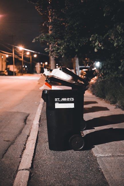 A black plastic wheelie bin with a white label and the words 'St. John's' printed on the front is positioned on a paved sidewalk next to a curb in an outdoor urban setting during nighttime. The bin is filled with various waste items, including cardboard boxes and black plastic bags, with some of the waste visibly sticking out from the open lid. The scene is illuminated by nearby streetlights, casting warm light and shadows on the pavement and surrounding foliage, which includes leafy trees and bushes along the sidewalk. In the background, there are additional streetlights, parked cars, and a glimpse of a quiet street, indicating a residential or suburban area. The image conveys an environment suitable for private rubbish collection or on-site waste disposal, which Rubbish Removal Swiss Cottage might support through their services in the NW3 area, emphasizing efficient and responsible rubbish management efforts.
